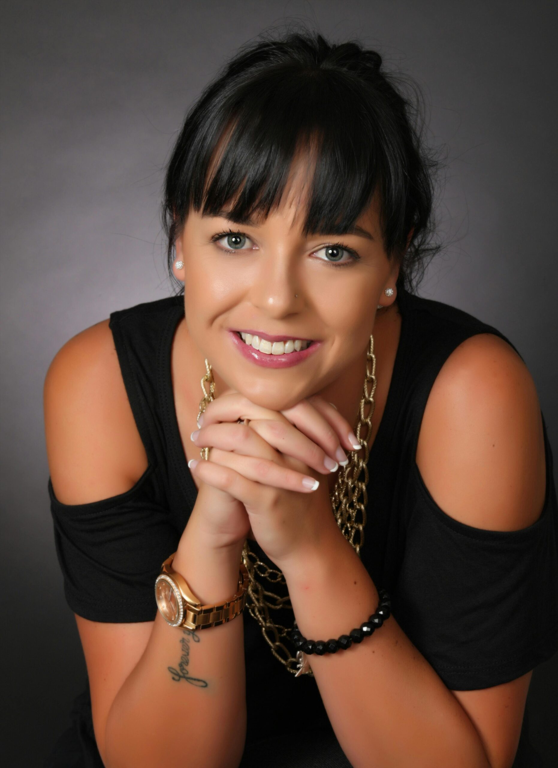 Portrait of an elegant woman smiling, showcasing jewelry and a tattoo against a dark background.