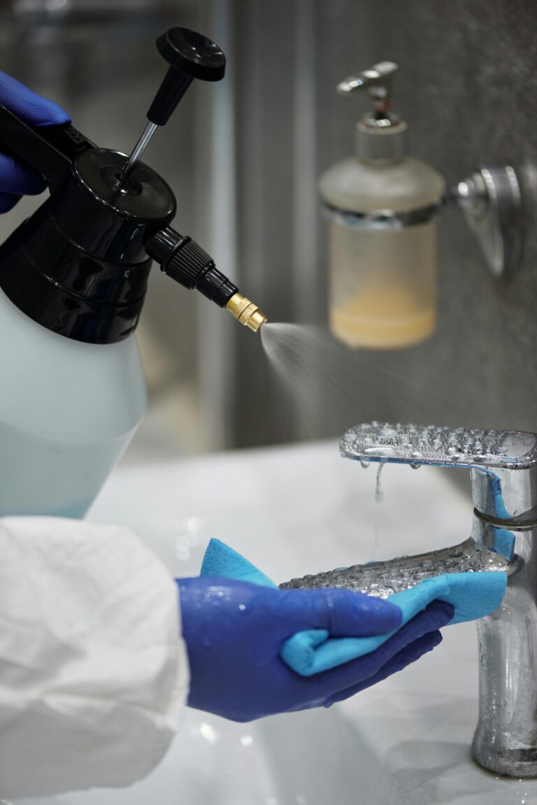 A person wearing gloves sprays and wipes a bathroom faucet for hygiene.